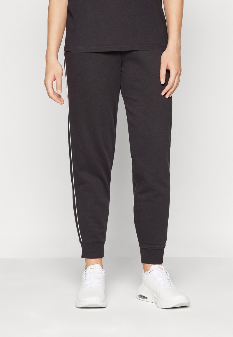 Calças de moletão pretas com riscas brancas nas laterais, punhos ajustados e um acabamento de tecido suave. Estilizadas com sapatilhas atléticas brancas.