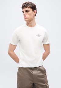 Νεαρός άνδρας που φοράει απλό λευκό T-shirt Calvin Klein και χακί παντελόνι cargo, στέκεται με τα χέρια στις τσέπες, κοιτάζοντας προς το πλάι.