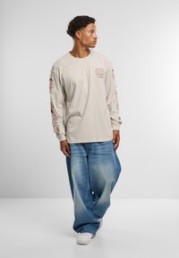 Beige langermet t-skjorte med røde grafiske design på ermene, sammen med vide, falmede blå jeans og hvite sneakers.