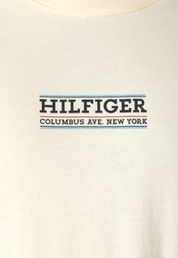 Krémová textilie s černým nápisem "HILFIGER" nad "COLUMBUS AVE. NEW YORK," orámováno tenkými modrými a červenými vodorovnými pruhy.