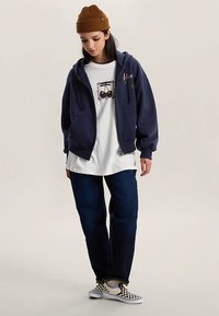 Marineblauer Hoodie mit Reißverschluss, weißes T-Shirt mit Kirschmotiv, dunkelblaue Jeans und schwarz-weiß kariertes Slip-On-Schuhwerk. Braune Strickmütze.