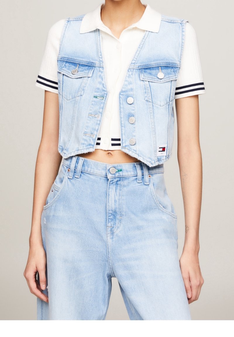 Ljusblå denimcroppad väst med fyra framfickor och silverdetaljer, parat med en vit ribbad skjorta med marinblå ränder.