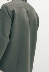 Grijze longsleeve shirt van glad, textuurachtig materiaal. Heeft een rugpas en een plooi voor bewegingsgemak. Eenvoudig ontwerp, zonder versieringen.