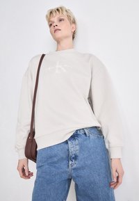 Light beige sweatshirt with oversized fit, ribbed cuffs, and a printed logo on the front, paired with light blue loose-fit jeans.