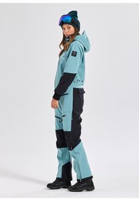 Skisuit in lichtblauw en zwart, met een capuchon, ritszakken en een slank ontwerp. Draag het met een zwarte beanie en kleurrijke goggles.