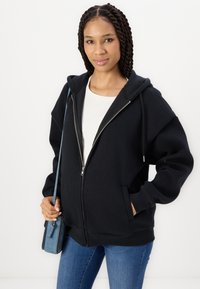 Saco preto com fecho éclair, de design espaçoso, com bolsos frontais e capuz forrado, combinado com uma camisa branca e jeans de ganga azul.