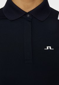 Marinblå kortärmad polo med ribbad krage, knappslå och en vit logopunkt på vänster sida. Slät, elastisk tyg.