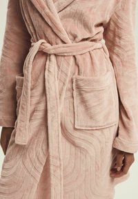 Jasnoróżowy pluszowy szlafrok o falistej teksturze, z paskiem wiązanym w talii, dwoma przednimi kieszeniami i kołnierzem szalowym. Miękki, przytulny materiał.