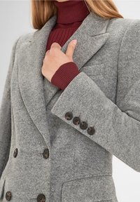 Blazer double boutonnage en mélangé de laine gris avec des épaules structurées, de larges revers et des boutons sombres. En dessous, un col roulé côtelé bordeaux.