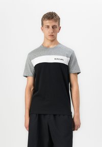 Ung mand iført en grå, hvid og sort Ripcurl farveblokket kortærmet skjorte og sorte bukser, stående imod en ensfarvet baggrund.