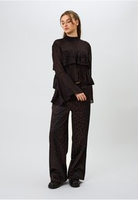 Melnas caurspīdīgas auduma drēbes ar slāņotiem volāniem, kurās ir augsts kakla izgriezums un platjūga bikses, abas ar tumšu leopardu rakstu.