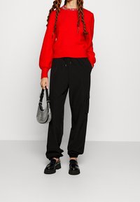 Nainen, joka on pukeutunut punaiseen villapaitaan, mustiin cargo-housuihin, mustiin niittisorjaisiin loafereihin ja pitää kädessään pientä hopeista käsilaukkua yksinkertaista taustaa vasten.