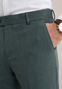 Szövetből készült, testre szabott zöld nadrág, lapos elsősorral, oldalsó zsebekkel és karcsú fazonnal.