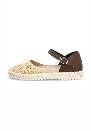 Zapato plano beige con diseño floral recortado, correa marrón en el tobillo con hebilla y suela blanca gruesa y texturizada.