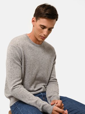 Maglione di lana grigio chiaro con collo rotondo, polsini e orlo a coste. Indossato con jeans di denim blu, seduto su uno sgabello di legno su uno sfondo bianco.