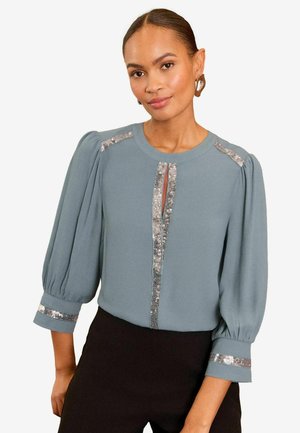 Blusa de color azul claro con mangas largas abullonadas. Presenta un corte en forma de llave y detalles de lentejuelas en el frente y los puños. Textura de tela suave.