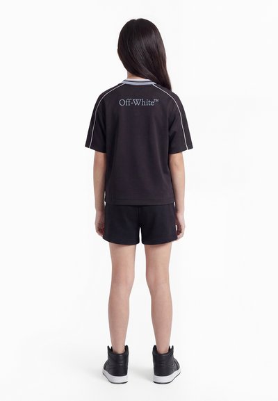 Κορίτσι με μακριά σκούρα μαλλιά στέκεται με την πλάτη γυρισμένη φορώντας μαύρο κοντομάνικο μπλουζάκι με το λογότυπο "Off-White", μαύρα σορτς και μαύρα ψηλά αθλητικά παπούτσια.