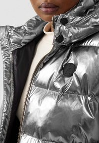 Giacca a piumino argentata metallica con zip frontale, collo alto e bottoni neri con logo in rilievo. Texture liscia e lucida con design trapuntato.