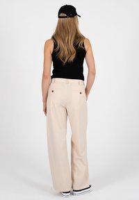 Femme portant un haut noir sans manches, un pantalon beige à jambes larges, une casquette noire et des baskets plateformes blanches, debout de dos.