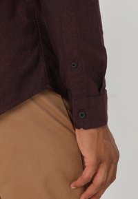 Primer plano de una persona que lleva una camisa de manga larga de color burdeos oscuro con puños abotonados y pantalones marrón claro, con la mano relajada a un lado.