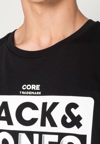 Černé bavlněné tričko s kulatým výstřihem, na kterém je bílé textové nápis "CORE TRADEMARK" a "BLACK & WHITE" v tučných kontrastních písmenech.