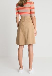 Falda de algodón color beige con forma acampanada, cintura alta y cierre de cremallera en la parte trasera. Acompañada de una blusa a rayas y zapatillas blancas.