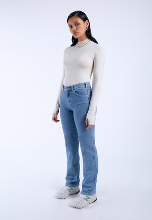 Getailleerde crème lange mouwen top, hoge taille lichte blauwe jeans en witte sneakers met donkere accenten, staand tegen een effen achtergrond.