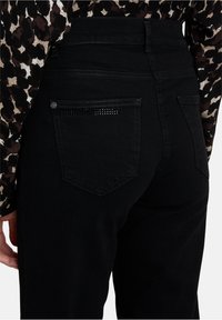 Close-up van een persoon die zwarte jeans draagt met strass details op de achterzak en een lang mouwendop met luipaardprint.