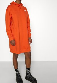 Personne portant une robe sweat à capuche longue orange avec une poche avant, des sandales noires à boucle et des chaussettes noires avec des lettres blanches sur un fond blanc.