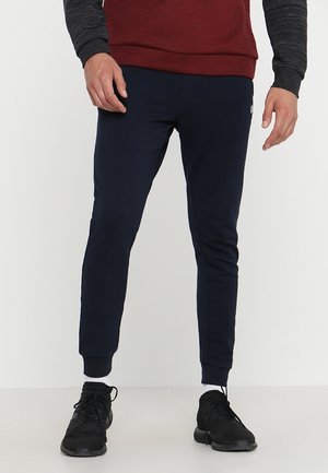 Mann trägt marineblaue Jogginghose, schwarze Sneakers, weiße Socken und ein rot-schwarzes Langarmshirt und steht vor einem schlichten Hintergrund.