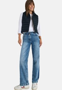Schwarze gepolsterte Weste über einem weißen Langarmtop, kombiniert mit weit geschnittenen, hellblauen Jeans und weißen Sneakers mit roten Akzenten.