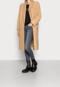 Henkilö, joka on pukeutunut beigeen polvipituiseen takkiin, mustaan paitaan, harmaisiin tiukkoihin farkkuihin ja mustiin paksupohjaisiin nilkkabotteihin, seisoo valkoista taustaa vasten.