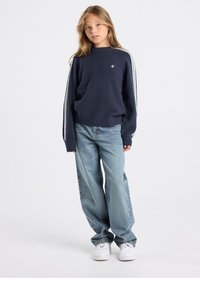 Pull en laine bleu marine avec des rayures blanches sur les côtés, col rond et ourlet côtelé, associé à un jean bleu clair ample et des baskets blanches.