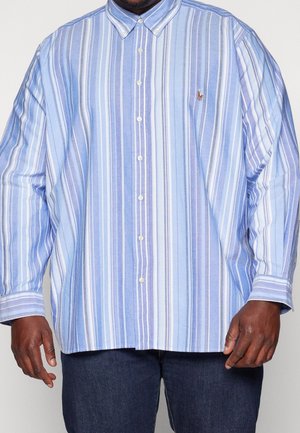 Mężczyzna ubrany w luźną koszulę na guziki z długim rękawem w pionowe niebiesko-białe paski, zestawioną z ciemnoniebieskimi dżinsami.