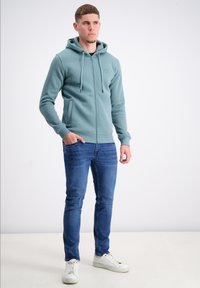 Gai zila jaku ar rāvējslēdzēju un priekšējo kabatu, savelkamu kapuci un ribotu aproci, apvienota ar tumši ziliem šauriem džinsiem un baltām tenisēm.