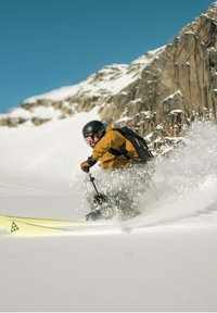 Eine Skifahrerfigur in einer leuchtend orangefarbenen Jacke und schwarzer Ausrüstung, mit einem schwarzen Helm und einer Skibrille, die durch den Schnee nahe einem felsigen Berg fährt.