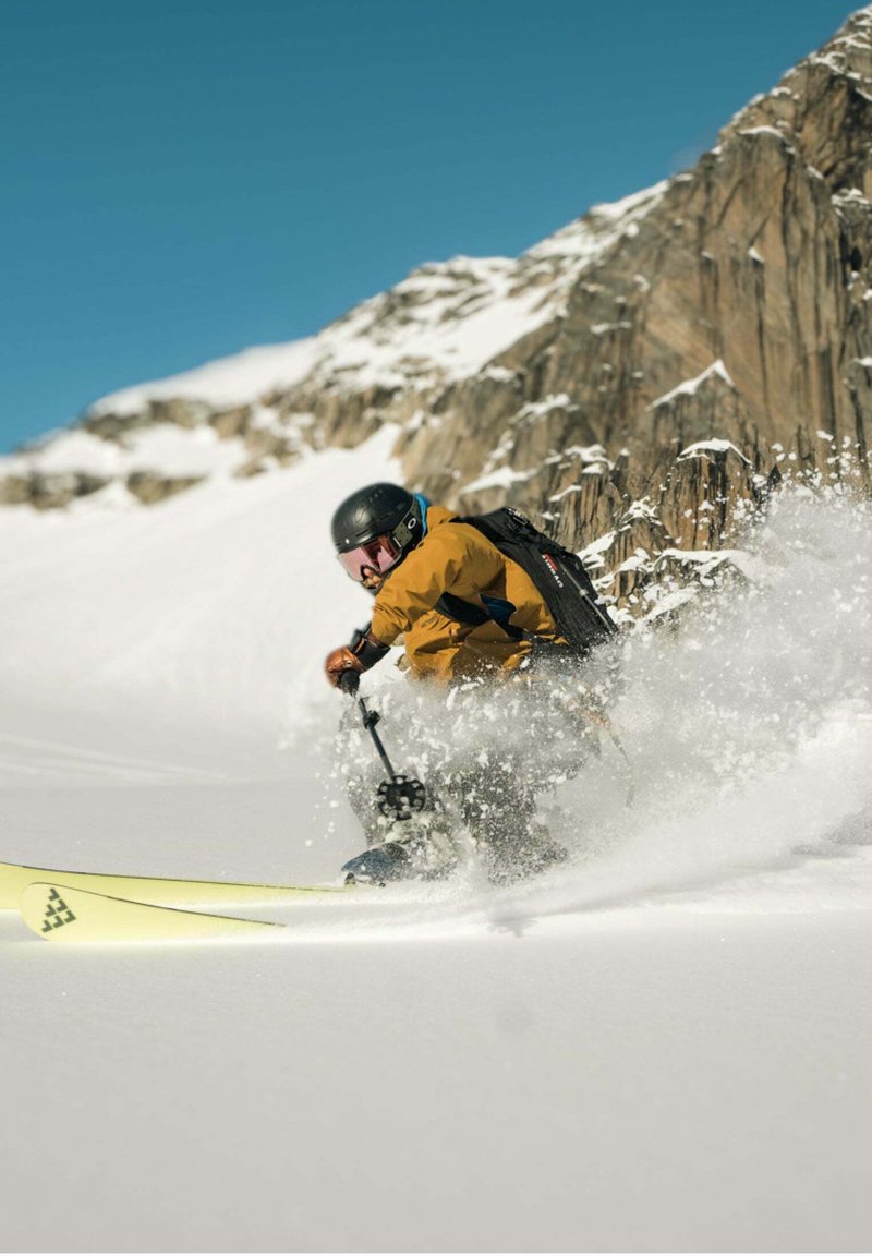 Eine Skifahrerfigur in einer leuchtend orangefarbenen Jacke und schwarzer Ausrüstung, mit einem schwarzen Helm und einer Skibrille, die durch den Schnee nahe einem felsigen Berg fährt.