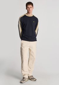 Sweatshirt bleu marine avec des rayures beiges et grises sur les manches, arborant un petit logo jaune. Associé à un pantalon cargo crème et des sneakers grises.