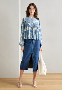 Lyseblå mønstrete bluse med lange ermer og samlet midje, kombinert med en høy midje denim blyantskjørt. Modellen holder en hvit tote bag.