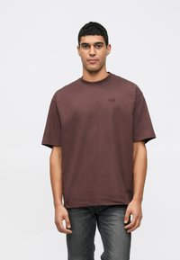 Mladý muž s krátkymi čiernymi vlasmi, oblečený v voľnom tmavočervenom tričku a vyblednutých čiernych džínsach, stojaci na pozadí s jednoduchým svetlým vzorom.