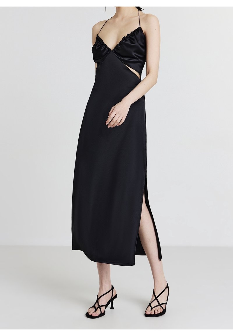 Robe en satin noir avec décolleté en V, corsage à volants, côtés ouverts et une fente haute. Caractérisée par des bretelles fines et une texture lisse et brillante.