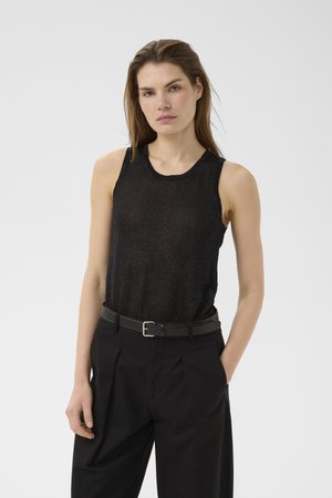 Femme portant un haut noir sans manches et un pantalon noir avec une ceinture noire, debout avec une main dans la poche, sur un fond uni.