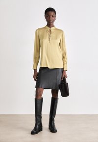 Blusa gialla a maniche lunghe con dettaglio al collo a fiocco, abbinata a una mini gonna di pelle nera e stivali neri sopra il ginocchio, mentre tiene una borsa scura.