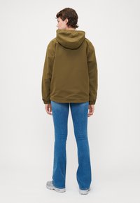 OLIVEN GRØNN HEKLET JAKKE MED HEKKE OG ELASTISKE MANSJETTER, BÆRT OVER LYS BLÅ FLAREDE BUKSER. STOFFET VIRKER GLATT MED EN MATTE FINISH.