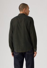 Camicia in denim verde scuro con maniche lunghe. Presenta un colletto appuntito, chiusure con bottoni e un orlo curvato. Cuciture visibili come dettagli.