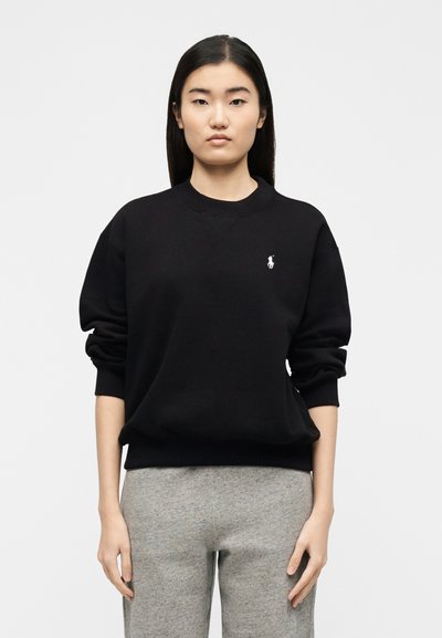 Γυναίκα με μακριά μαύρα μαλλιά που φοράει μαύρο φούτερ με λευκό λογότυπο και γκρι παντελόνι, στέκεται μπροστά σε απλό λευκό φόντο.