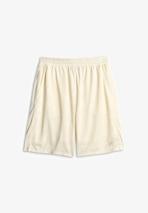 Femme vêtue d'une tenue de sport blanche sans manches, de baskets blanches, de chaussettes et d'une casquette blanche, posant avec une main dans la poche et l'autre ajustant la casquette.