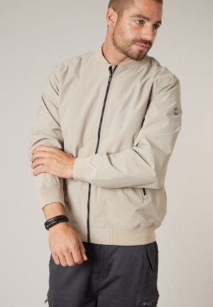Homme portant une veste zippée beige clair et un pantalon foncé ajuste sa manche gauche, debout devant un fond neutre uni.