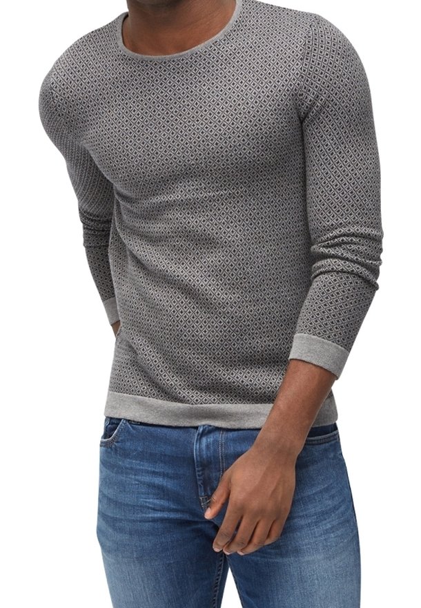 Homme portant un pull gris ajusté à manches longues avec un motif en losange et un jean bleu, main reposant près de la poche.