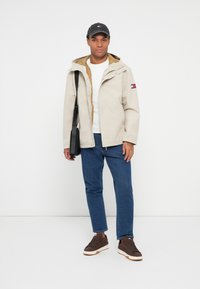 Hombre vestido con chaqueta beige, camisa blanca, jeans azules, zapatillas marrones, gorra negra y llevando una bolsa de hombro negra, de pie contra un fondo blanco.